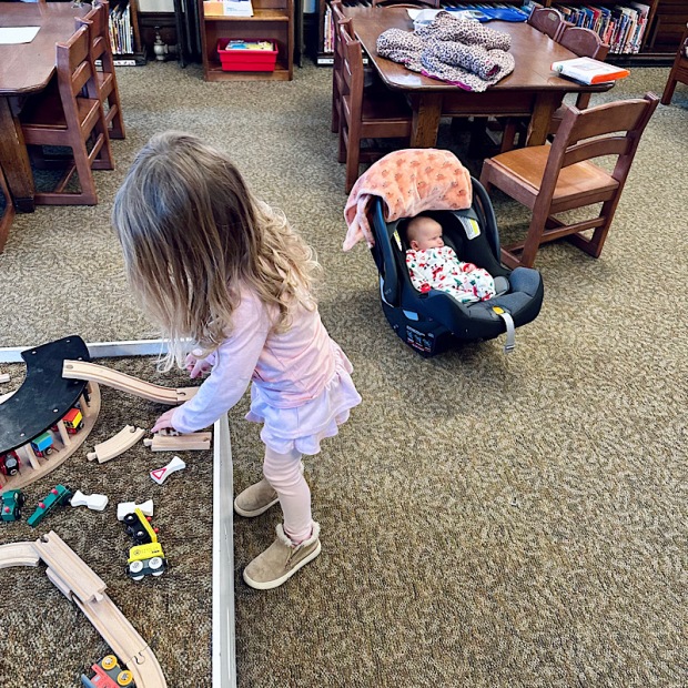 Girl playing with train set at the library while baby rests in carseat
